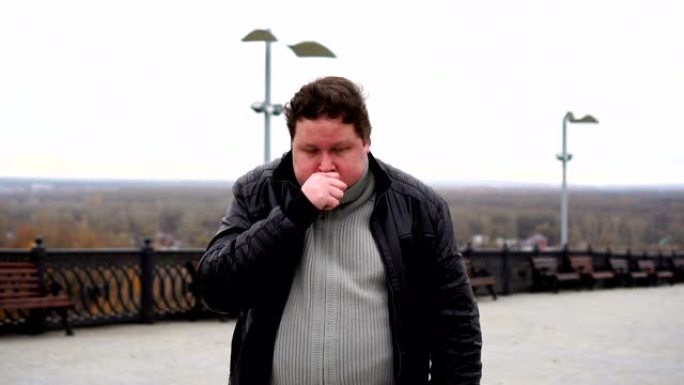 在寒冷的秋天天气中，放大胖子看着相机并在户外咳嗽的肖像。