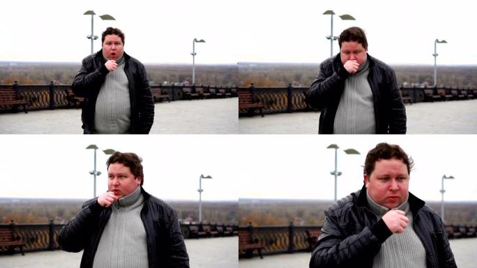 在寒冷的秋天天气中，放大胖子看着相机并在户外咳嗽的肖像。
