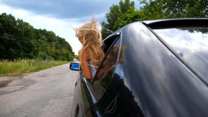 在阴天的天气里，小女孩斜靠在车窗外。小孩把头伸出汽车，长长的金发随风飘扬。关闭后视图
