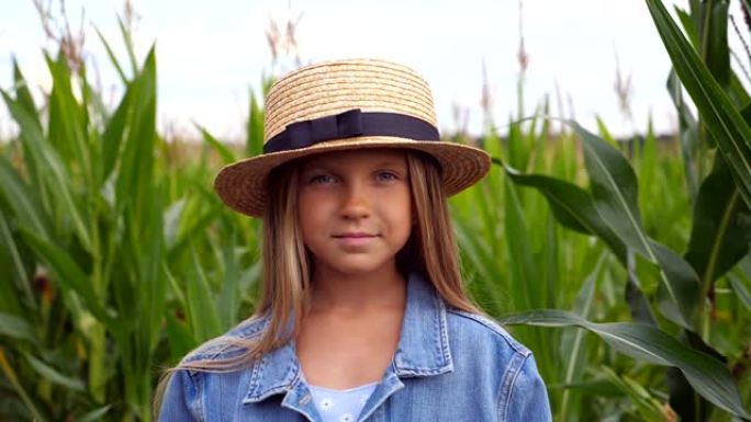 快乐的小孩的肖像，长长的金发站在玉米田模糊的背景下。戴着草帽的微笑小女孩在有机农场的草地上看着相机。