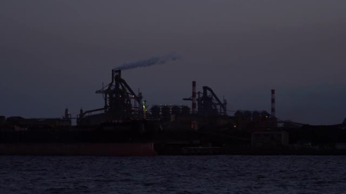 工业船在煤电厂前的码头卸煤，而烟雾在夜间通过烟囱排出