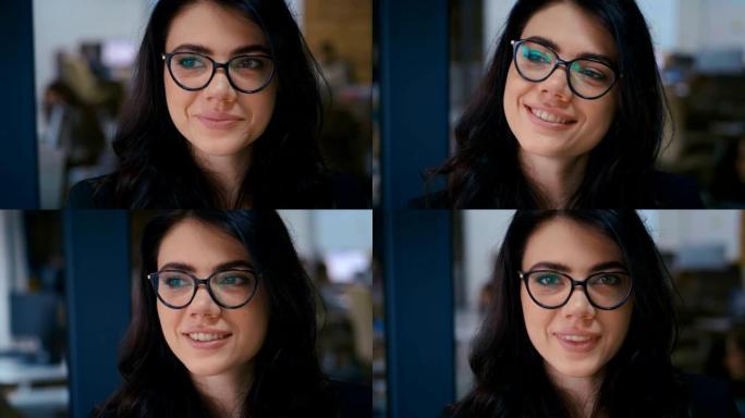 美丽微笑自信的年轻女子漂亮的脸看着办公室里的相机，特写肖像。戴眼镜。背景办公室工作。