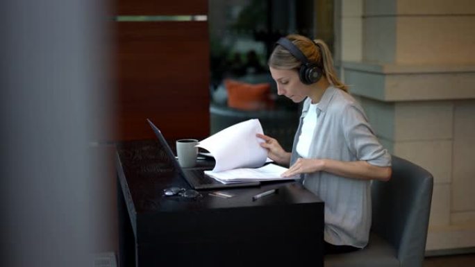 蓝牙耳机中的智能女企业家坐在笔记本电脑的台式机上，从纸质报告中检查信息