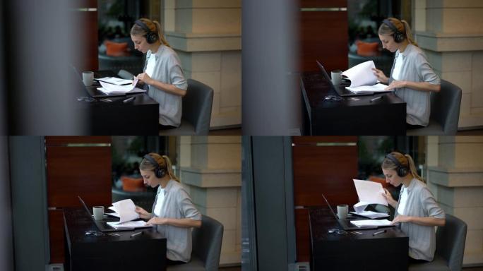 蓝牙耳机中的智能女企业家坐在笔记本电脑的台式机上，从纸质报告中检查信息