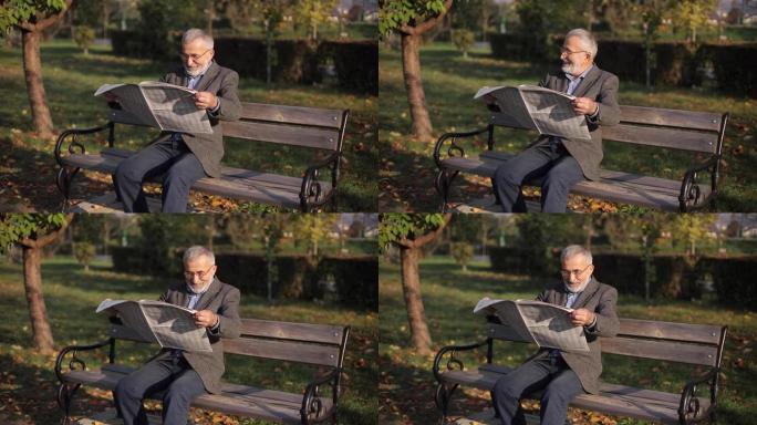 穿着灰色夹克留着漂亮胡须的英俊祖父坐在公园的长椅上看报纸。戴眼镜的白发老人