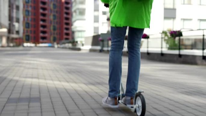 后视图跟随绿色制服的年轻食品快递员在城市街道上骑着踏板车，背着隔热背包，向上倾斜