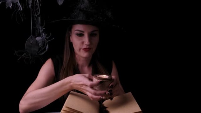 迷人的神秘女巫在黑色背景上有着鲜红的嘴唇，看着一个魔法球，同时在一本咒语书旁边施放咒语