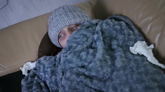 覆盖着灰色格子的年轻白人男子在帽子和围巾冻僵在家里感觉寒冷，生病的男性有发热流感温度症状包裹在毯子里