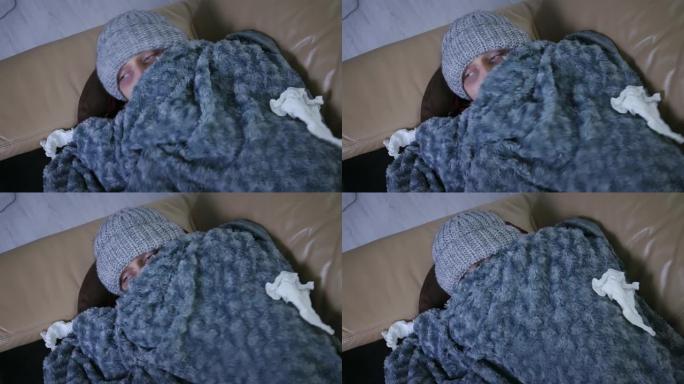 覆盖着灰色格子的年轻白人男子在帽子和围巾冻僵在家里感觉寒冷，生病的男性有发热流感温度症状包裹在毯子里