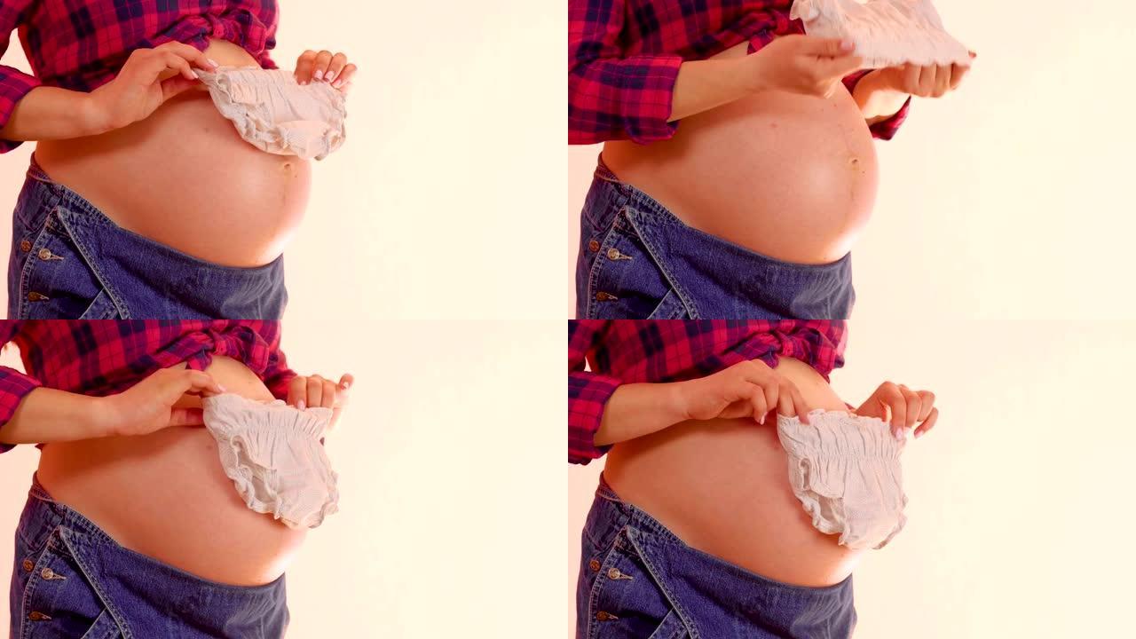 孕妇手里拿着新生儿衣服在她裸露的大肚子上。孕妇拿着可爱的婴儿衣服。怀孕和孕产概念。中枪