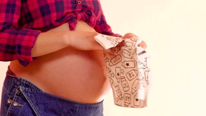 孕妇手里拿着滑块在她裸露的大肚子上。孕妇拿着可爱的婴儿衣服。怀孕和孕产概念。中枪