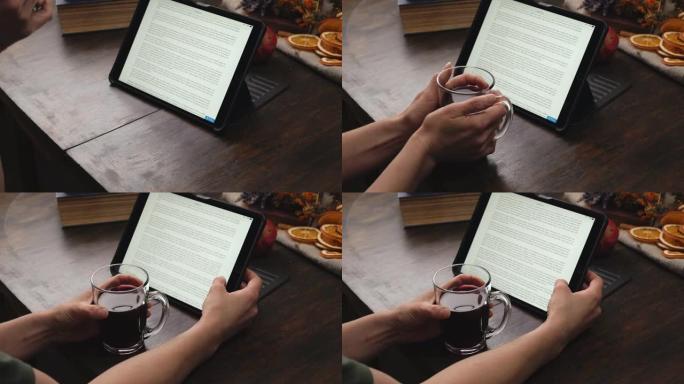 女人双手拿着装有热酒的杯子，在平板电脑上看书。