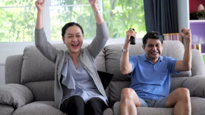 成熟的亚洲夫妇在电视节目中欢呼体育，兴奋的情绪在家里的客厅沙发上，人们和娱乐场所。