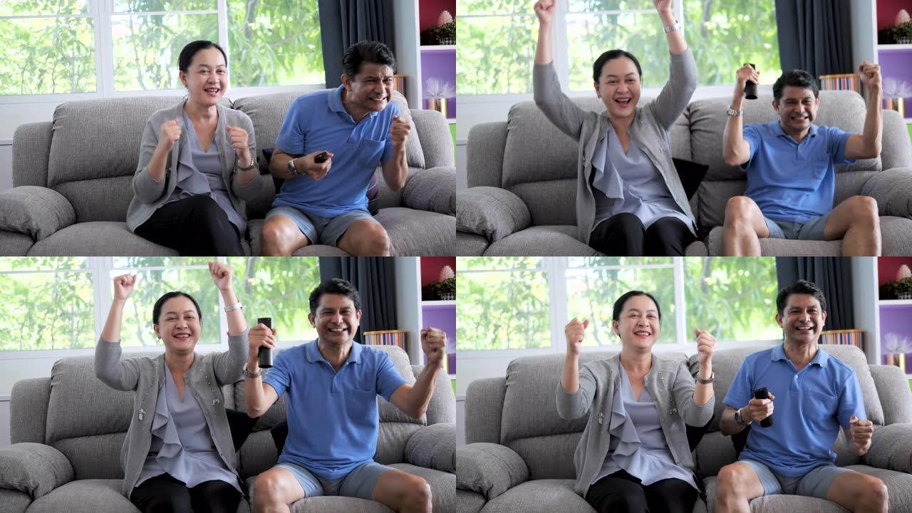 成熟的亚洲夫妇在电视节目中欢呼体育，兴奋的情绪在家里的客厅沙发上，人们和娱乐场所。