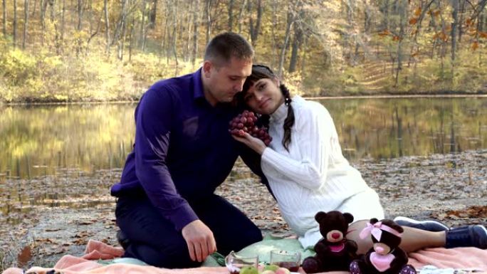 在阳光明媚的秋天，一个怀孕的女孩和一个男人在池塘附近的大自然中与一束红葡萄坐在一起摆姿势。他们拥抱并