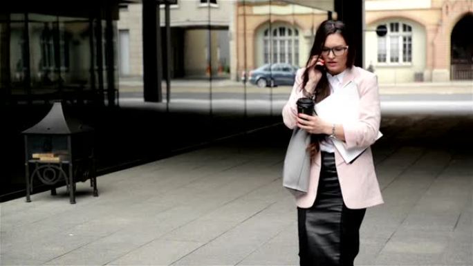 一位年轻的戴着眼镜的压力很大的女商人带着文件穿过城市，并通过电话交谈。她放下论文以承受压力