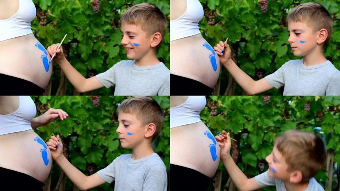 微笑的小男孩用刷子画在他怀孕的妈妈婴儿的脚印的肚子上。怀孕概念。特写