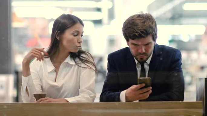 商人在手机上工作，而沮丧的无聊女孩在购物中心咖啡馆里坐在他旁边争吵