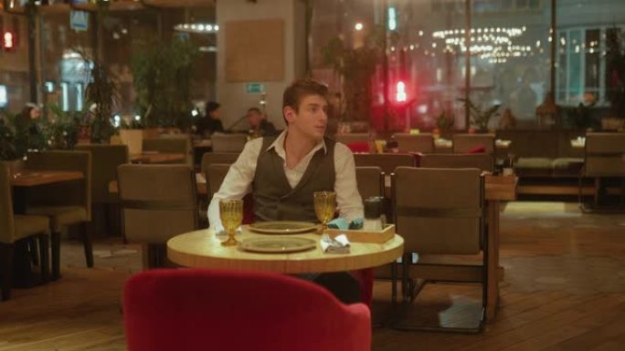 英俊的年轻人独自坐在一家时尚的餐厅里环顾四周。男人希望他的女朋友在餐馆里。摄像机从上到下布线。