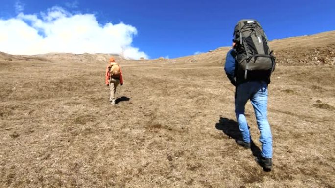 两名游客摄影师留着胡子的男人和女孩戴着帽子和太阳镜的背包，在雪山的背景下，手里拿着相机，在黄色的草地