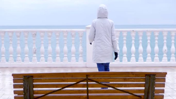 穿着白色夹克和兜帽的女人在美丽的露台上，可以看到海滨的海景。后视图。