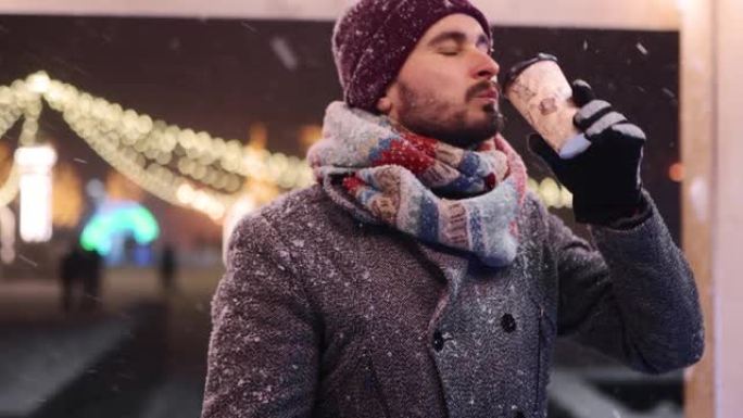 穿着灰色外套和针织围巾的大胡子时尚男人从纸杯里喝咖啡，对着镜头微笑。背景圣诞市场和新年冬季交易会。下