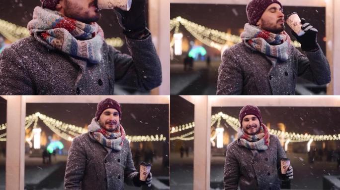 穿着灰色外套和针织围巾的大胡子时尚男人从纸杯里喝咖啡，对着镜头微笑。背景圣诞市场和新年冬季交易会。下