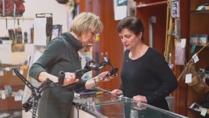 迷人的金发成熟，50岁的女人在小狩猎商店检查弩，在妇女销售人员的协助下。
