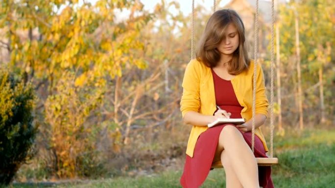 美丽的沉思女孩坐在花园里的秋千上，在笔记本上写字，一位浪漫的年轻女子在秋天自然，概念生活方式和艺术，