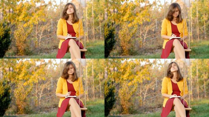 美丽的沉思女孩坐在花园里的秋千上，在笔记本上写字，一位浪漫的年轻女子在秋天自然，概念生活方式和艺术，