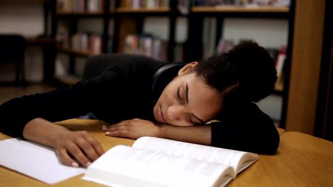 一个穿着黑色毛衣的美国黑人女学生，累了，在现代图书馆里的桌子上睡着了，桌子上有书籍和笔记。教育、人、