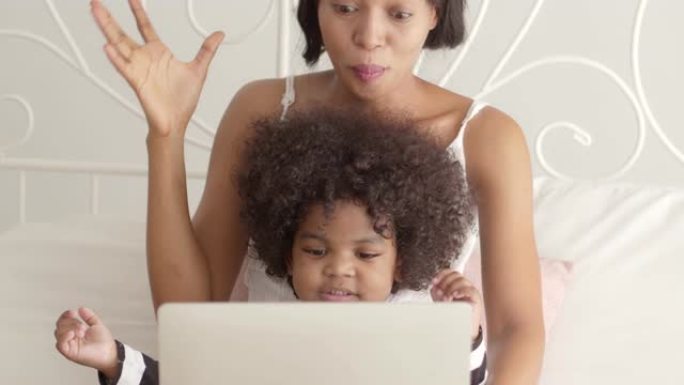 年轻的非洲母亲在笔记本电脑上看卡通或有趣的歌曲，她的儿子在白色床上。慢动作镜头。