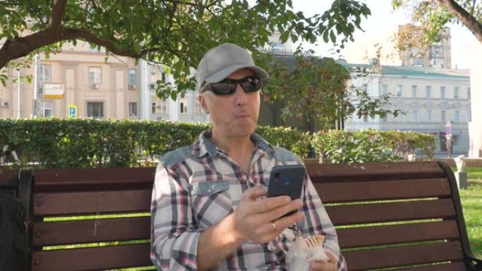 戴墨镜的白人男子坐在公园的长凳上吃智能手机的点击