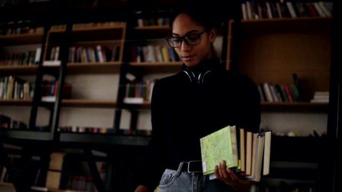 开朗的非洲妇女站在小图书馆里走下楼梯，手里拿着一堆书。穿着黑色毛衣，脖子上戴着耳机，戴着时尚的眼镜。