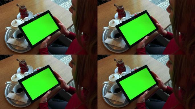 一位金发女子坐在咖啡馆用餐的餐桌旁，看着一块绿屏的平板电脑-从上方特写