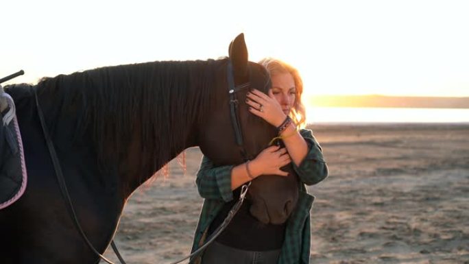 金发女人抚摸着拥抱的马。黑色种马享受日落的女士