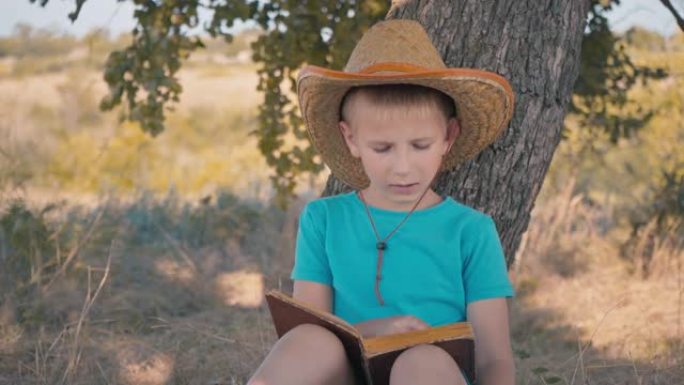 一个男孩坐在树下看书。一个戴着草帽的孩子在大热天读书。暑假。在树荫下休息，手里拿着一本书。快乐的童年