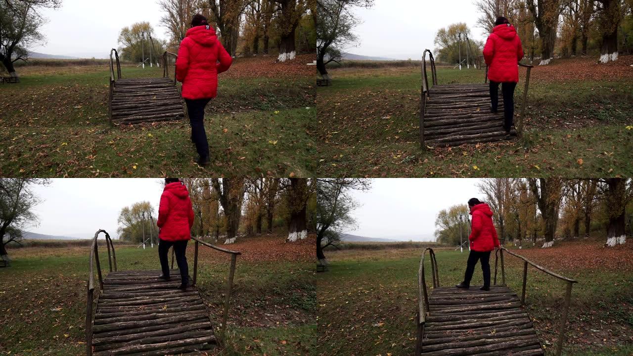 一名身穿红色外套的妇女在秋季公园的木桥上行走
