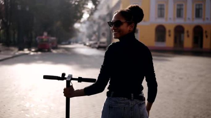 mix竞速女子在城市街道上行走的罕见镜头。穿着黑色外套和太阳镜的女孩在早晨的城市与电动踏板车一起行走
