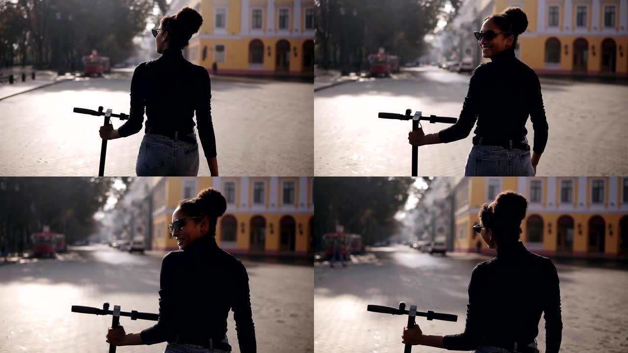 mix竞速女子在城市街道上行走的罕见镜头。穿着黑色外套和太阳镜的女孩在早晨的城市与电动踏板车一起行走