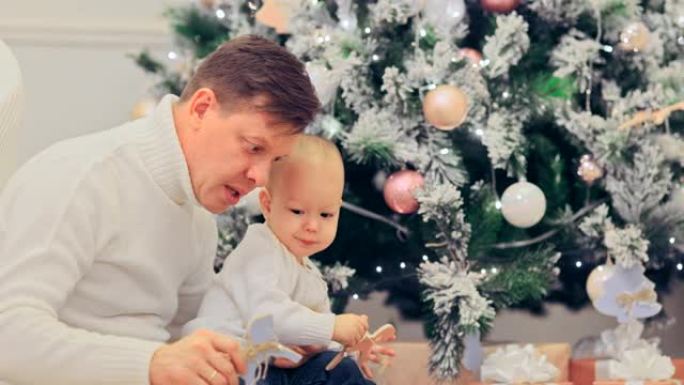 爸爸玩把婴儿抱在膝盖上，和他玩木马。快乐的童年。带玩具的圣诞树旁舒适的氛围。