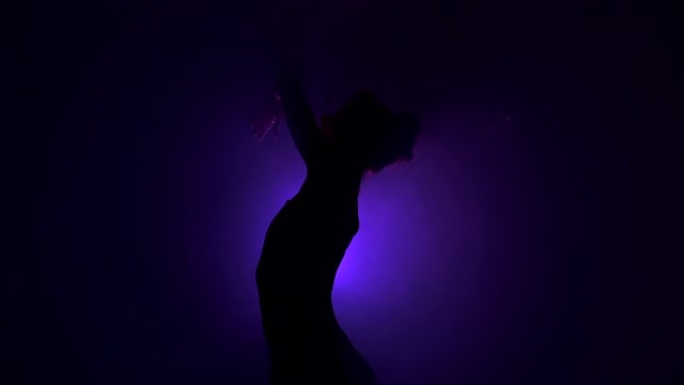 年轻美丽的芭蕾舞演员在烟舞台上跳舞现代芭蕾舞。用手在紫罗兰色的聚光灯背景下进行平稳的运动。现场穿着黑