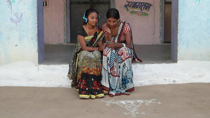 两名妇女在印度拉贾斯坦邦农村的家中戴着耳机听音乐，穿着传统服装，色彩鲜艳，印花艺术设计，当地工艺纱丽