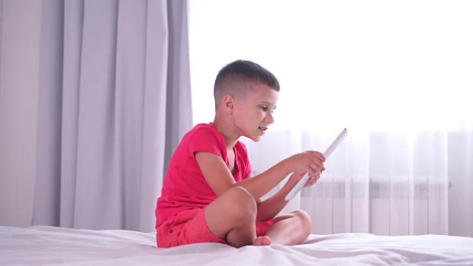 活跃的男孩坐在床上，在家玩平板电脑