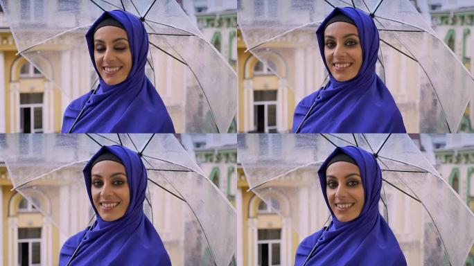 年轻迷人的穆斯林妇女戴着头巾，拿着雨伞，看着相机，在雨中微笑，在背景中建造