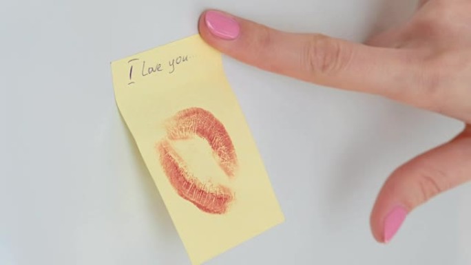 特写女人的手用吻粘在纸条上，在白色的墙上写下 “我爱你”。