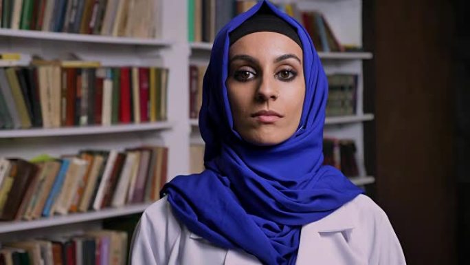 年轻美丽的穆斯林女子戴着头巾站在图书馆里，表情严肃地看着镜头