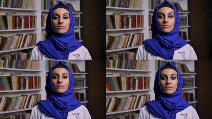 年轻美丽的穆斯林女子戴着头巾站在图书馆里，表情严肃地看着镜头