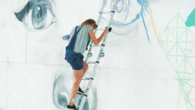 穿着牛仔工作服的女孩完成了绘画，从高楼梯上下来。美丽的年轻金发女人在城市街道墙上用气溶胶喷雾涂鸦大眼