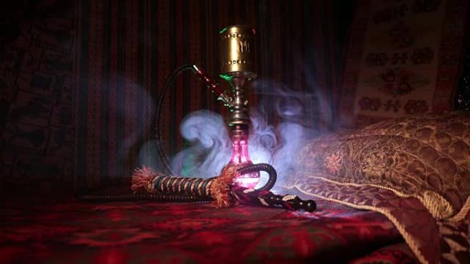 水烟碗上的水烟热煤在阿拉伯内部制造蒸汽云。地毯上的东方装饰品。黑暗中带背光的时尚东方水烟。滑块镜头。
